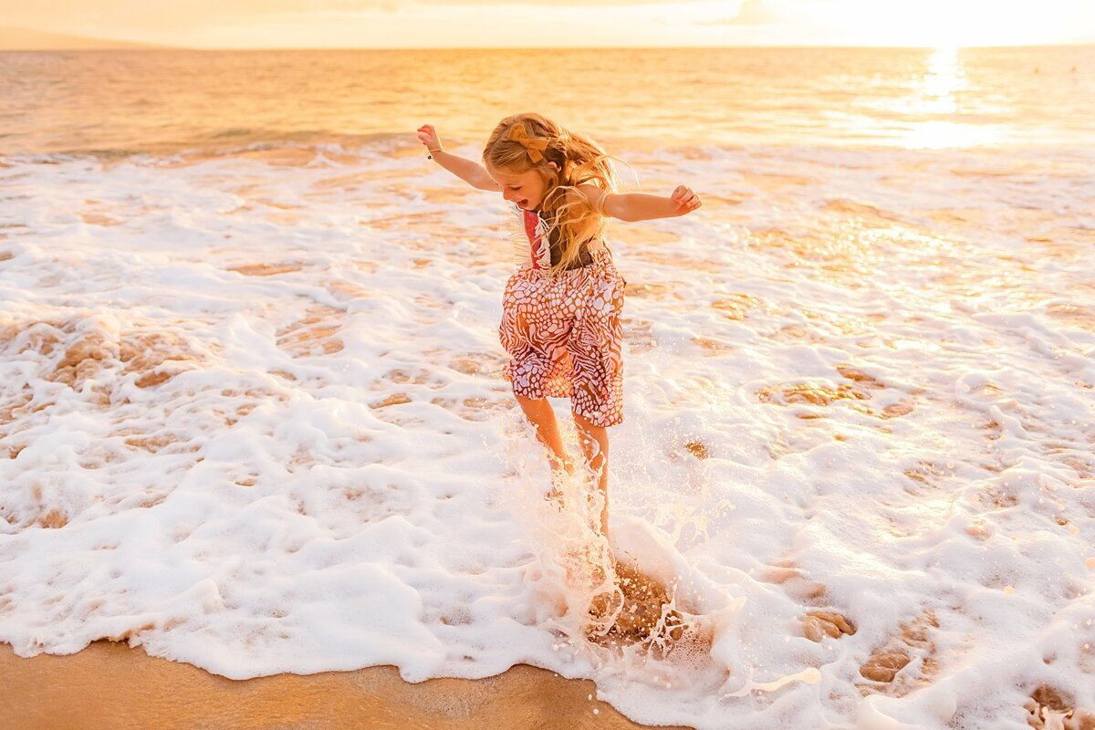 Girl lifts her hands in the air while jumping over a crashing wave at the beach on Maui for her photoshoot at sunset