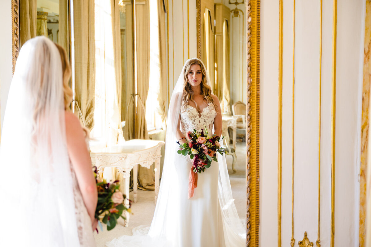 Bride looking in mirror in bridal suite at gosfield hall with  autumn bouquet