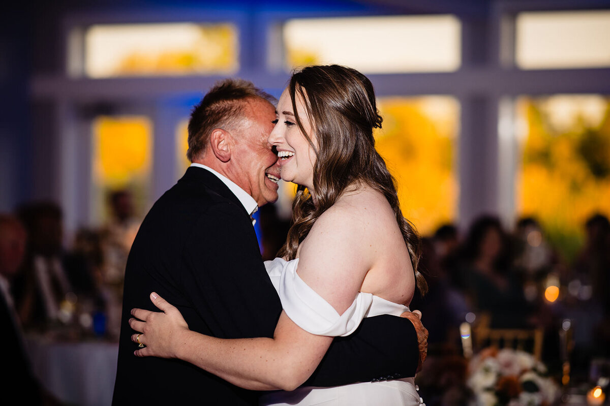 A bride in a white off-the-shoulder dress shares a dance and a laugh with a man in a black suit