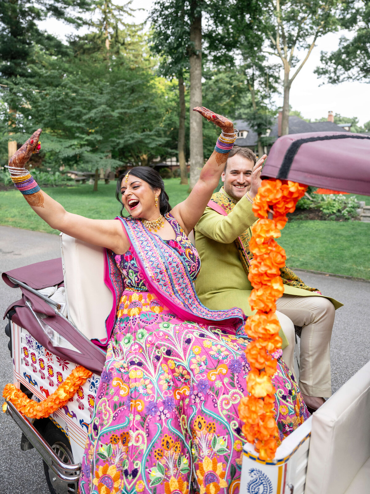 A joyful couple dressed in colorful traditional attire rides a decorated rickshaw adorned with orange flowers. They are smiling and waving, surrounded by greenery. The woman wears a vibrant dress and the man wears a light green outfit.