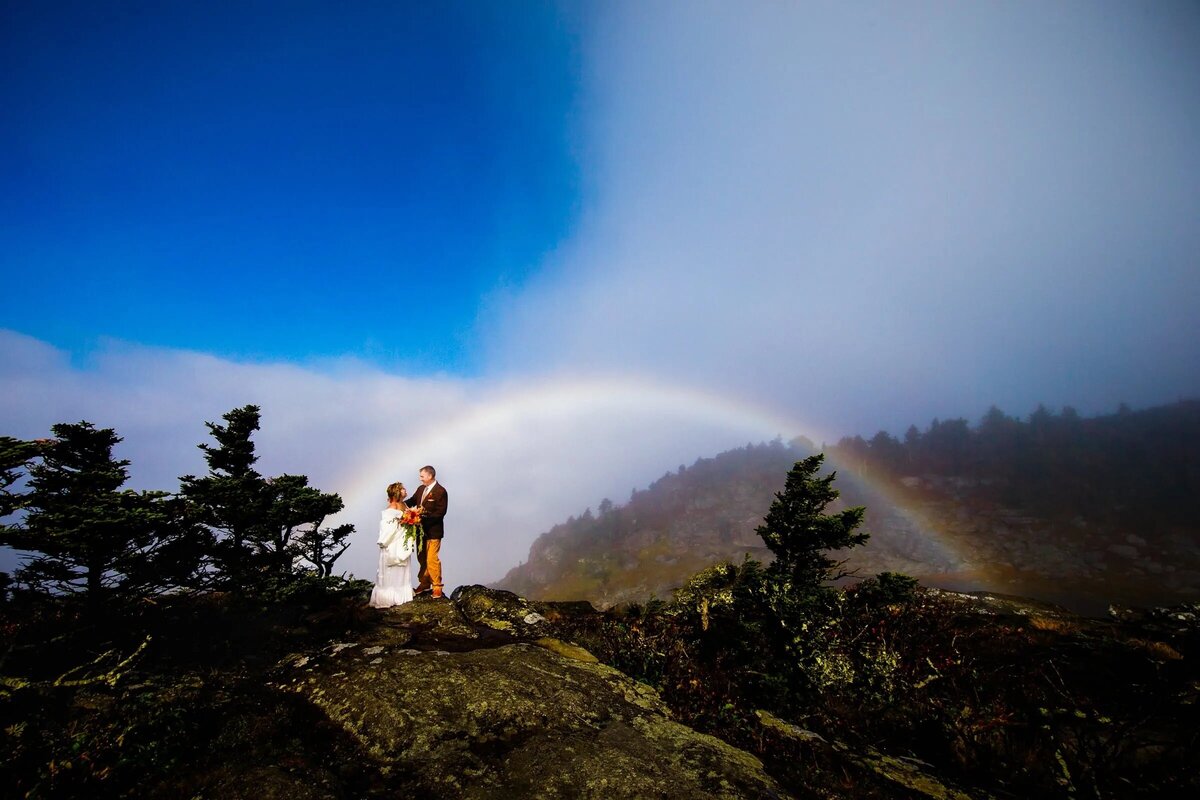 A bride and groom stands poised in a mountain setting, adjusting his cufflink, with a faint rainbow and mist in the background