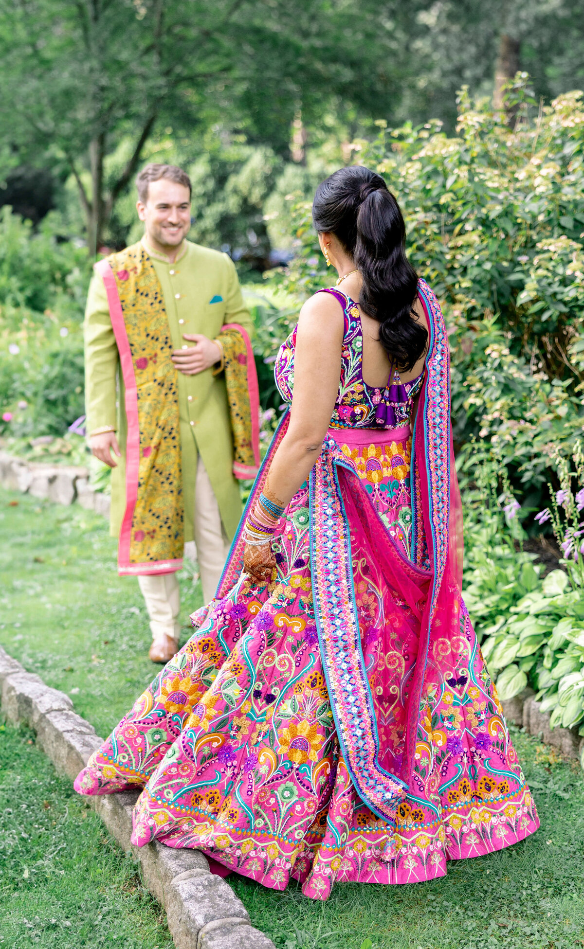 A woman in a vibrant, traditional Indian dress adorned with intricate patterns walks on grass in a garden, facing a man in a green sherwani with a colorful dupatta. They are surrounded by lush greenery and blooming flowers.