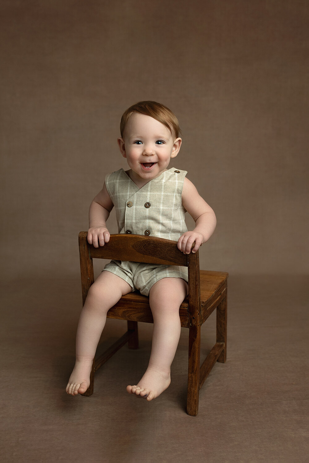 A happy toddler boy sits backwards on a wooden chair in a studio in a green onesie