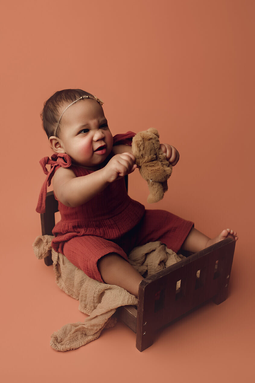 a one year old girl holding a teddy bear on a wooden bed srunching up her face