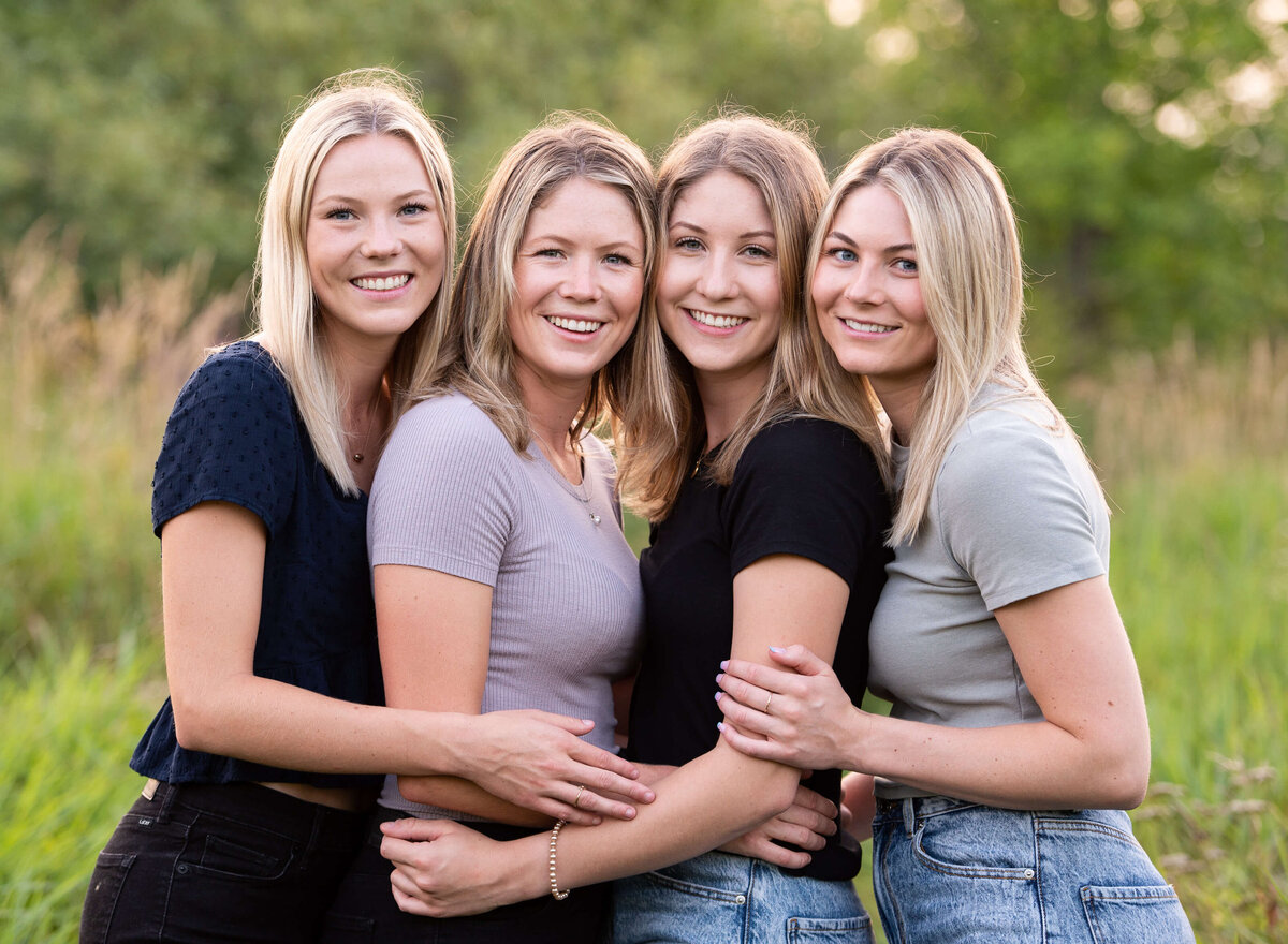 four adult sisters hug and smile together in a grassy field at sunset golden hour.  Taken by Ottawa family photographer JEMMAN Photography