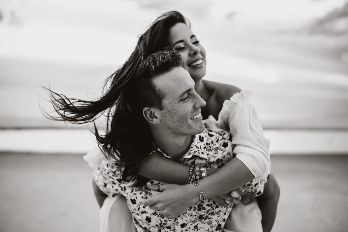 a black and white portrait of a mom and dad having fun doing piggy back rides on the beach