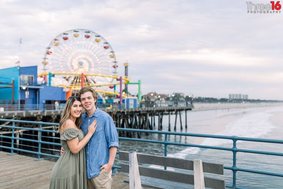 Engaged couple cozy up as they pose for engagement photos on the Santa Monica Pier with the Ferris Wheel in the background