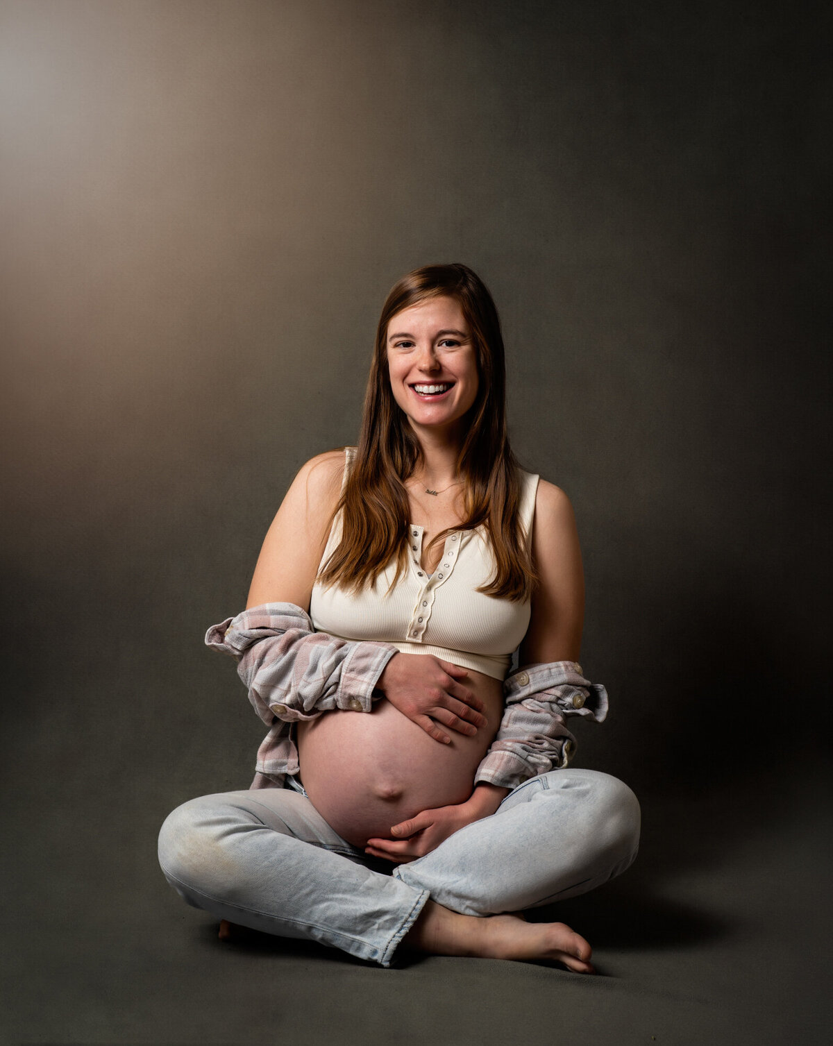 A mama to be sits criss cross apple sauce and holds her belly while smiling during her portrait session  with an Asheville Maternity Photographer