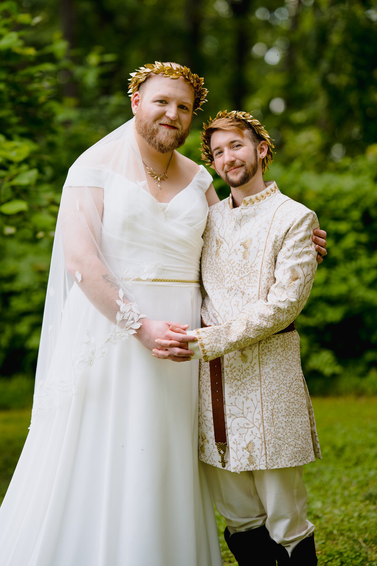 A wedding couple with one arm around each other and the other in front holding hands. They are smiling and standing in a small wooded area.