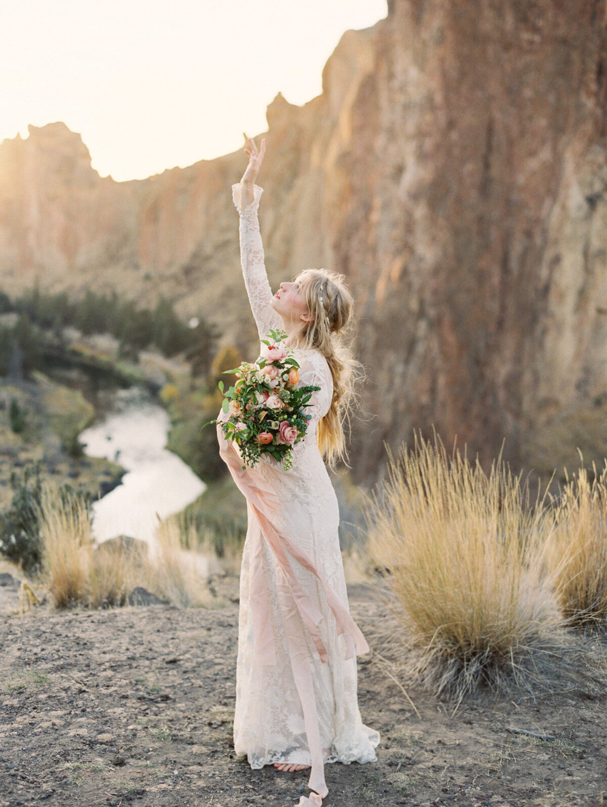 A bride in light pink wedding attire holds a bouquet and raises her arm to the sky in a canyon.