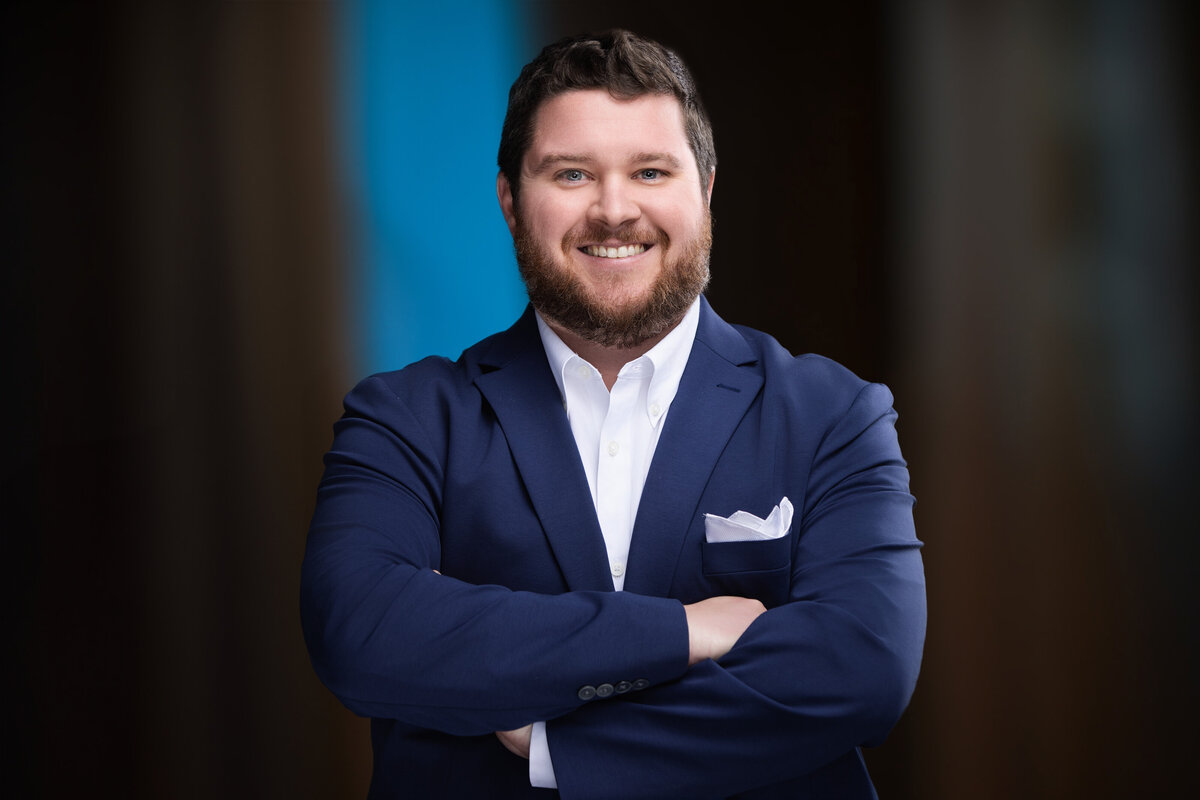 This professional headshot features a bearded man with neatly styled dark hair, exuding confidence with a warm smile. He is dressed in a sharp navy blue suit paired with a crisp white dress shirt and a neatly folded white pocket square. His arms are crossed, projecting a strong, approachable demeanor. The blurred background with a hint of blue adds a professional and modern touch to the composition. Taken in Cincinnati, this headshot is ideal for corporate and personal branding, emphasizing professionalism and approachability.
