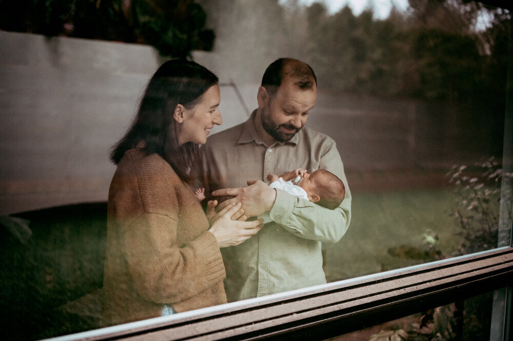 Candid newborn session captured through a window—parents smiling down at baby, highlighting quiet connection during a lifestyle session in Portland.