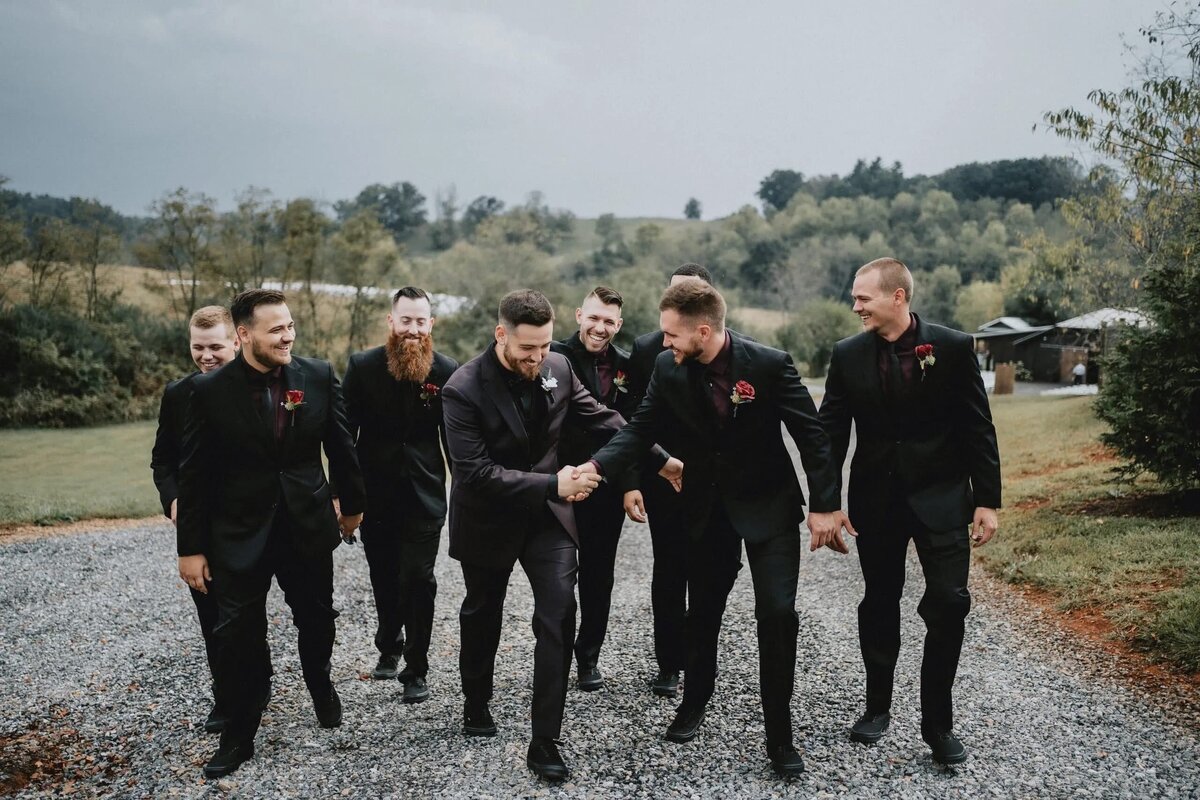 A line of groomsmen walking down a gravel path, laughing and adjusting their attire,