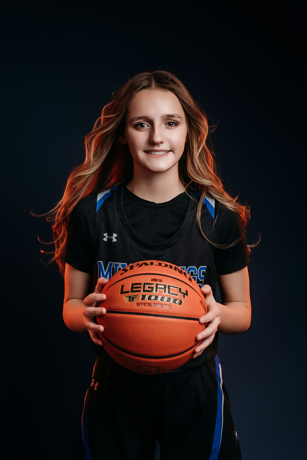 A teenage girl in a sports jersey holds a basketball in front of a blue solid backdrop illuminated by an orange glow around her for her senior graduation photoshoot, captured by a fine art studio photographer in Bemidji, Minnesota
