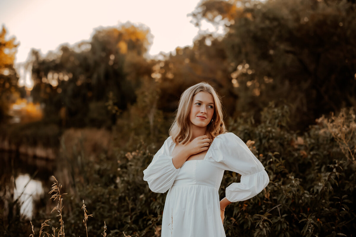 high school girl senior photos in a white dress with a pond in the background during golden hour