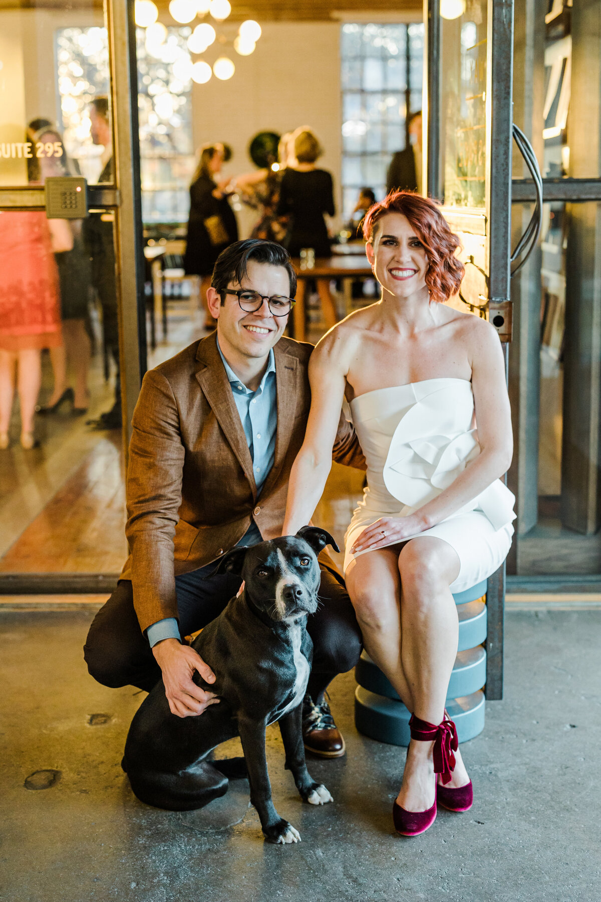 Portrait of a bride and groom with their dog on their wedding day in Dallas, Texas. The bride sits on a stool on the right and is wearing a sleeveless, simple yet elegant, white dress with red heels. The groom is kneeling down on the left and is wearing a brown blazer with dark dress pants. Their dog sits in front of the groom and is mostly black with a white stripe running down their face and chest.