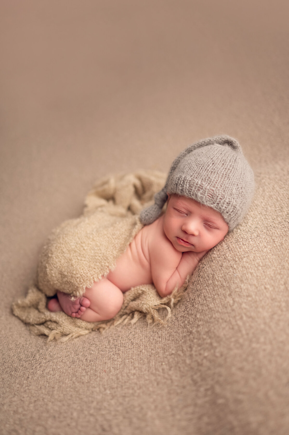 Portrait of a sleeping infant laying on a fuzzy beige background wearing a coordinating neutral hat and blanket.