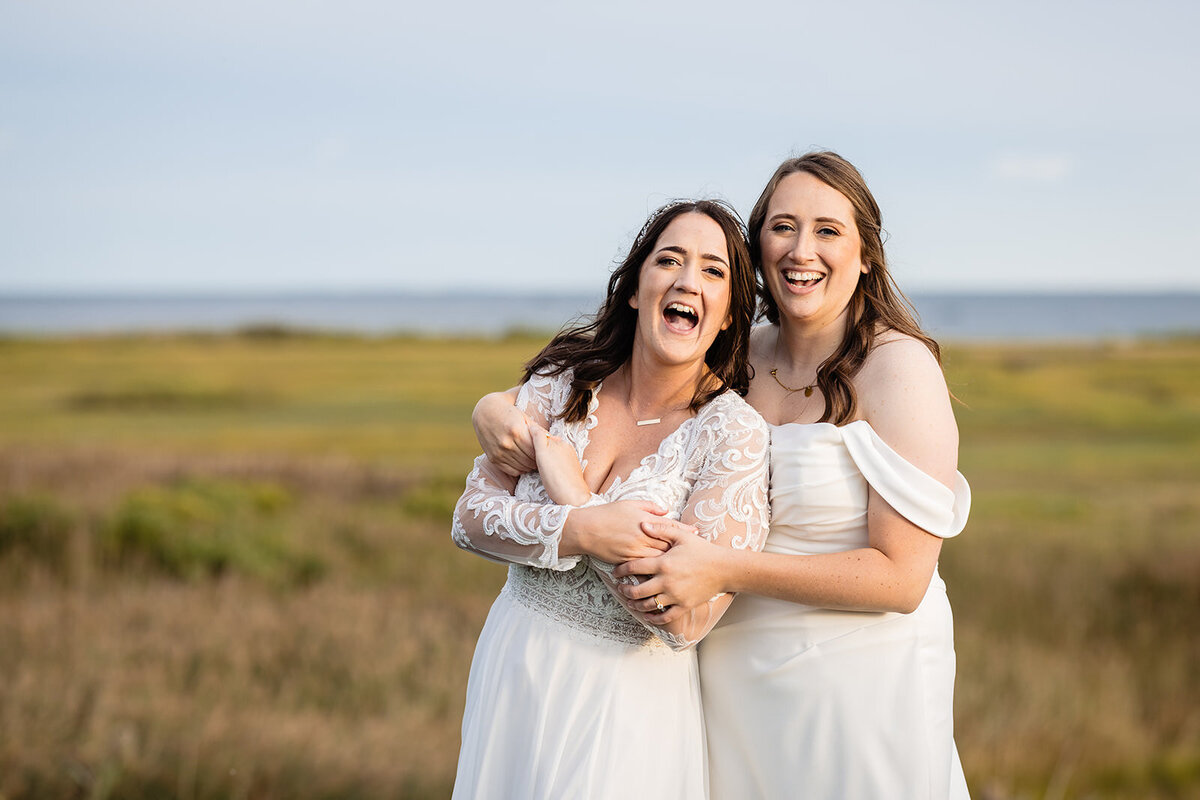 The two brides embrace and laugh, standing in a grassy field with a water view and blue sky behind them.