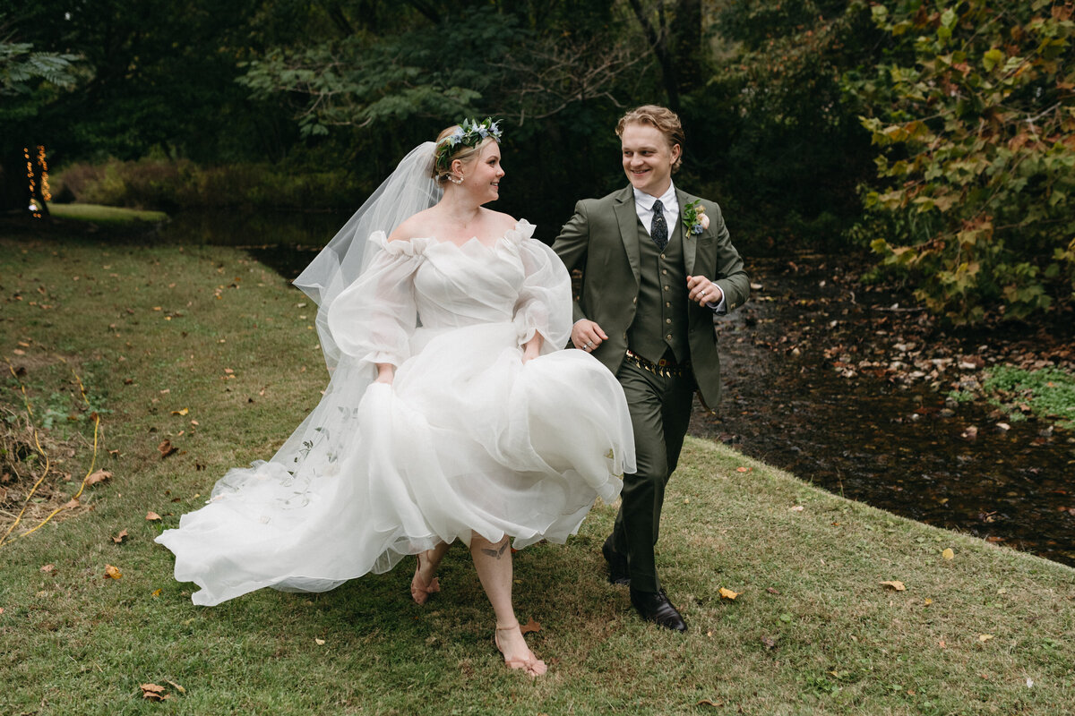 Bride and groom running hand-in-hand across a grassy meadow beside a creek, the bride lifting her flowing gown and veil embroidered with greenery, capturing a playful, whimsical moment from their garden-inspired wedding day.