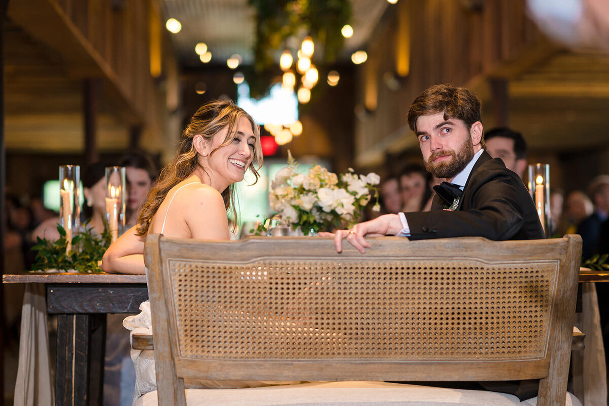 A bride and groom seated at their wedding table, smiling and looking back towards the camera.