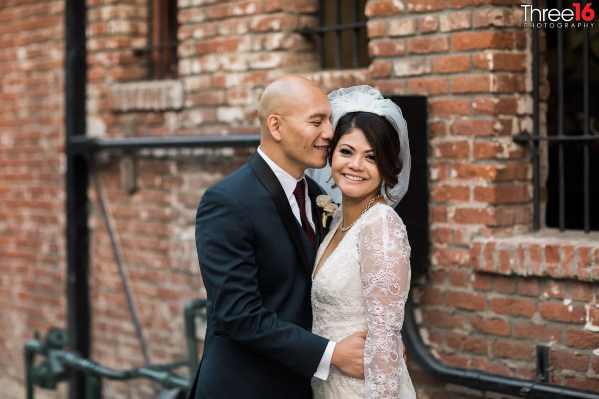Groom whispers into his Bride's ear as she smiles for the camera in front of a brick building in a Fullerton alleyway