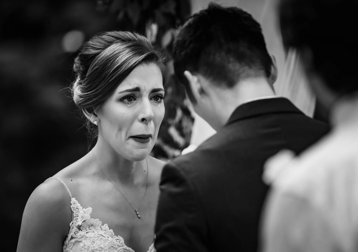 A poignant black and white photo capturing a bride's emotional expression during a wedding ceremony, with the groom facing away from the camera.