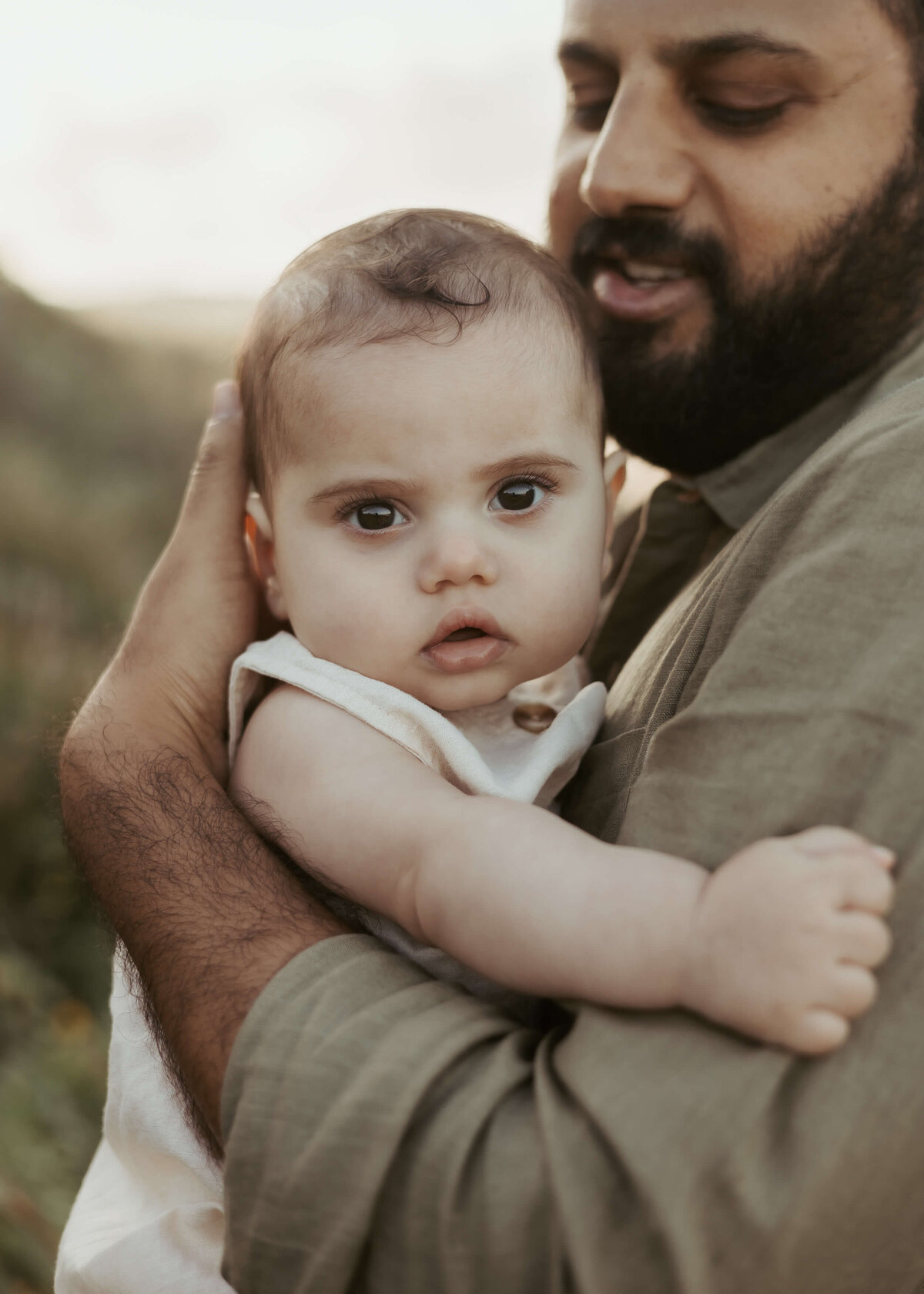 A beautiful baby staring into the camera while his dad's arms.