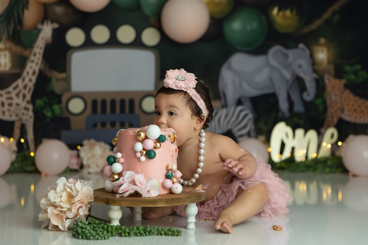 A toddler girl in pears and a pink tutu leans over for a bite of a pink cake in a jungle themed studio