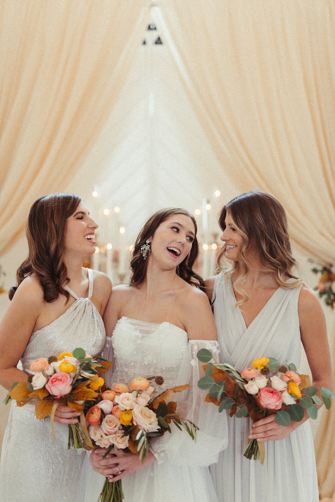 A bride wearing a white wedding gown poses smiling with bridesmaids wearing light gray gowns with bouquets in hand.