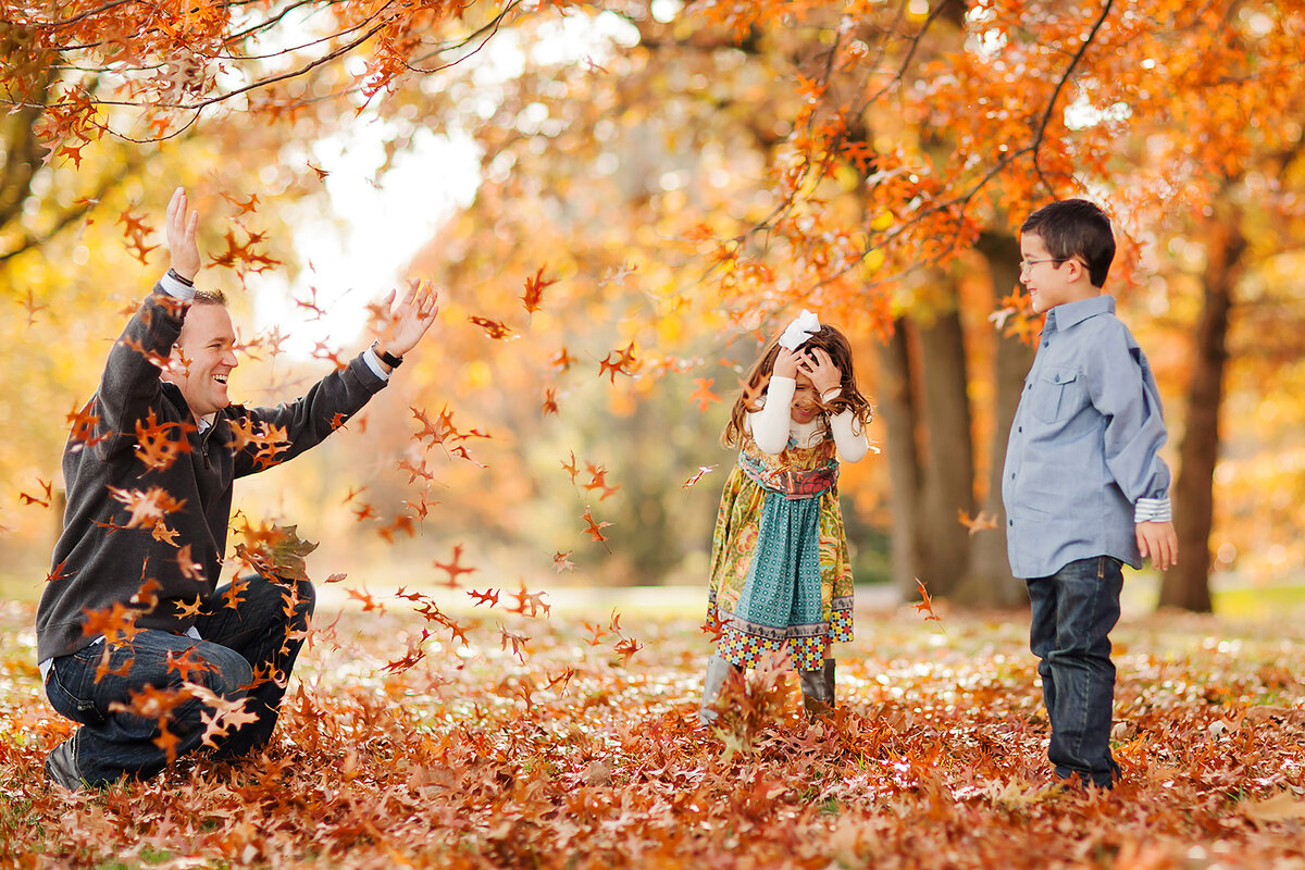 Dad is bent down throwing fall leaves in the air while his two kids stand nearby. His daughter is covering her face.