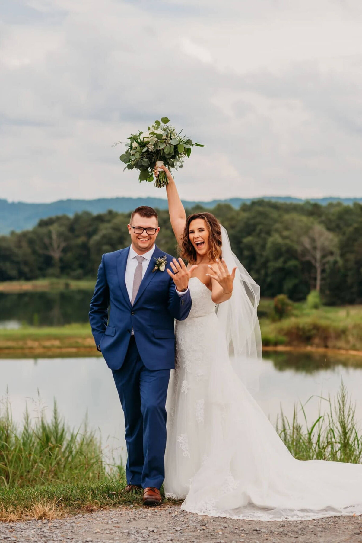 Photo of a bride and groom standing side-by-side and cheering as they show off their wedding rings and stand in front of a pond where the Mountain View's