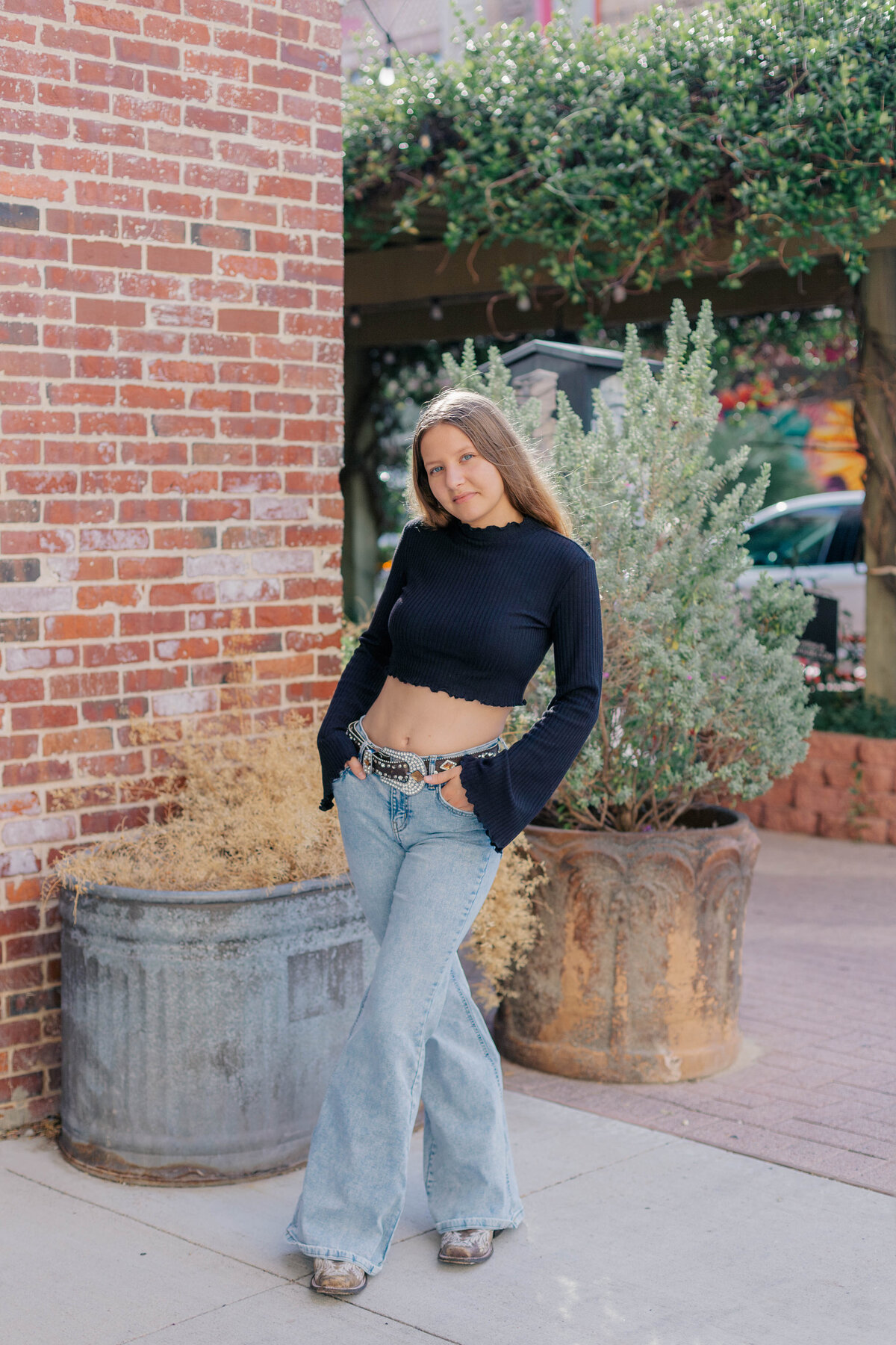 A young girl stands on the sidewalk in front of a coffee house patio while wearing a black long sleeve crop top with jeans a belt and cowboy boots