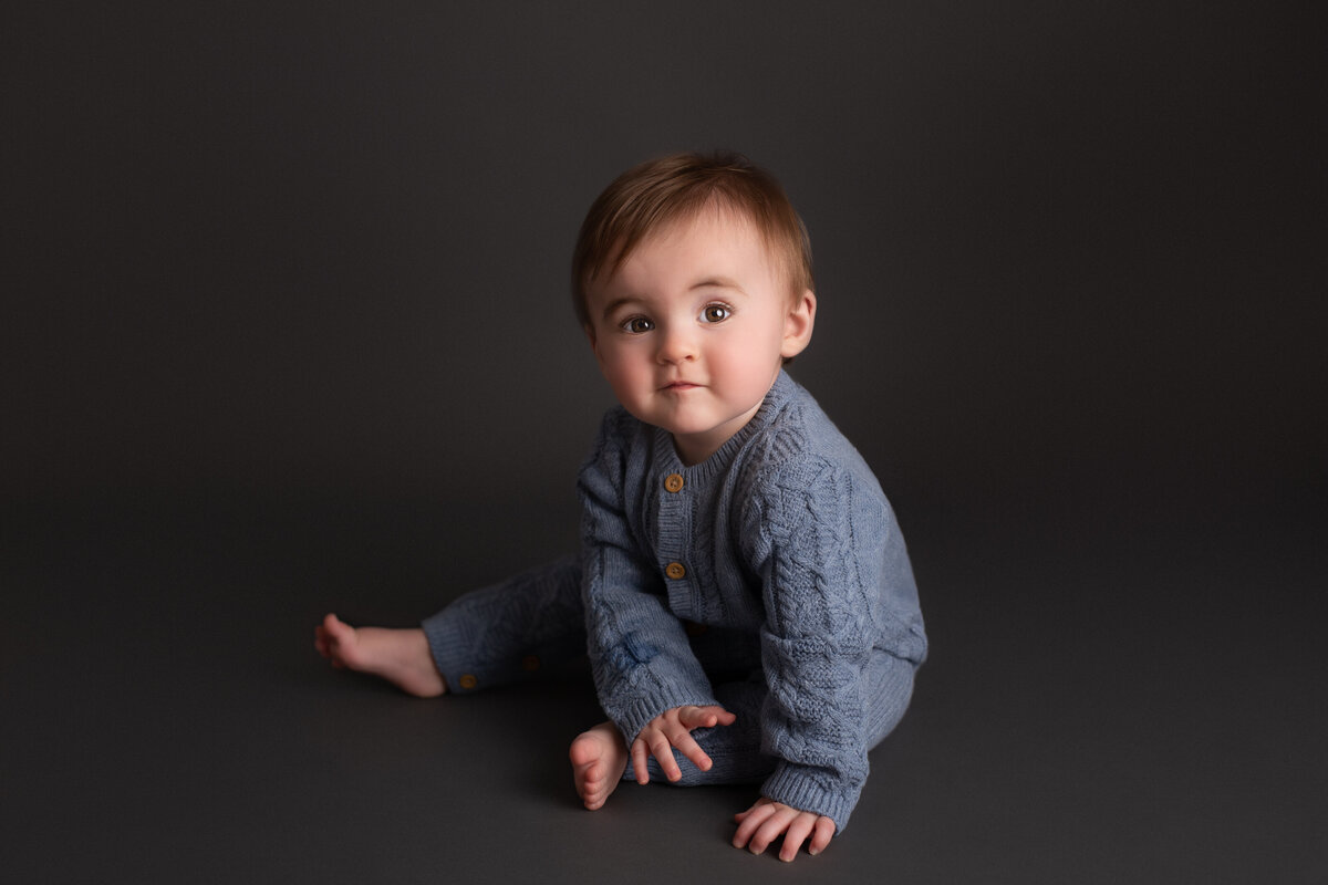 Baby boy dressed in a blue knitted romper sitting on a grey backdrop.
