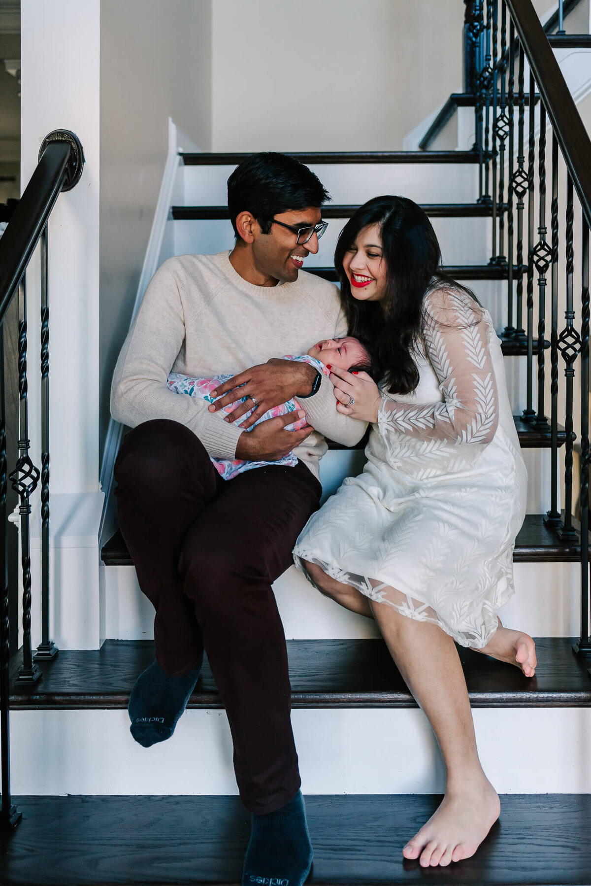newborn photo of parents with a sleeping baby girl laughing on the stairs in their Northern VA home