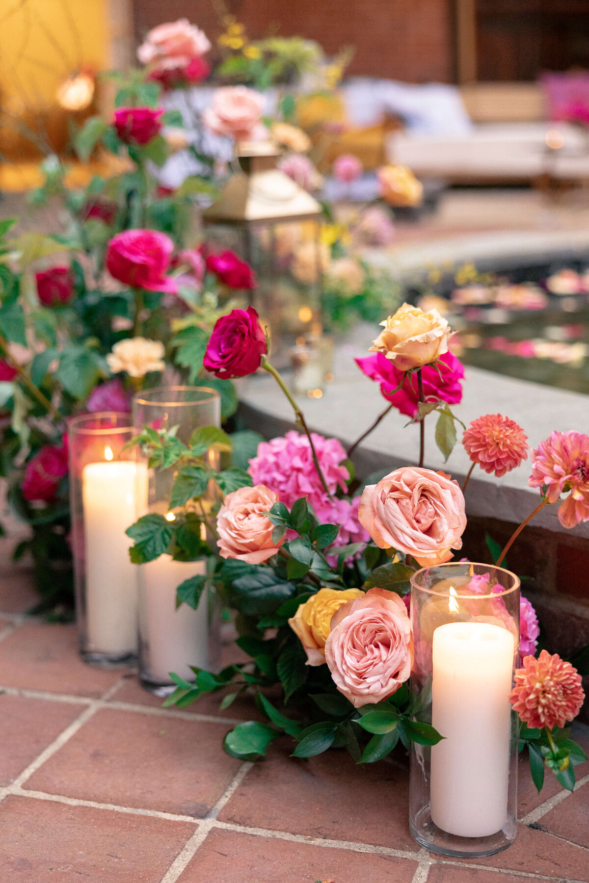 A decorative arrangement of pink and red roses intertwined with green leaves surrounds lit candles in glass holders. The setting appears to be near a pool or water feature, creating an elegant and romantic atmosphere.