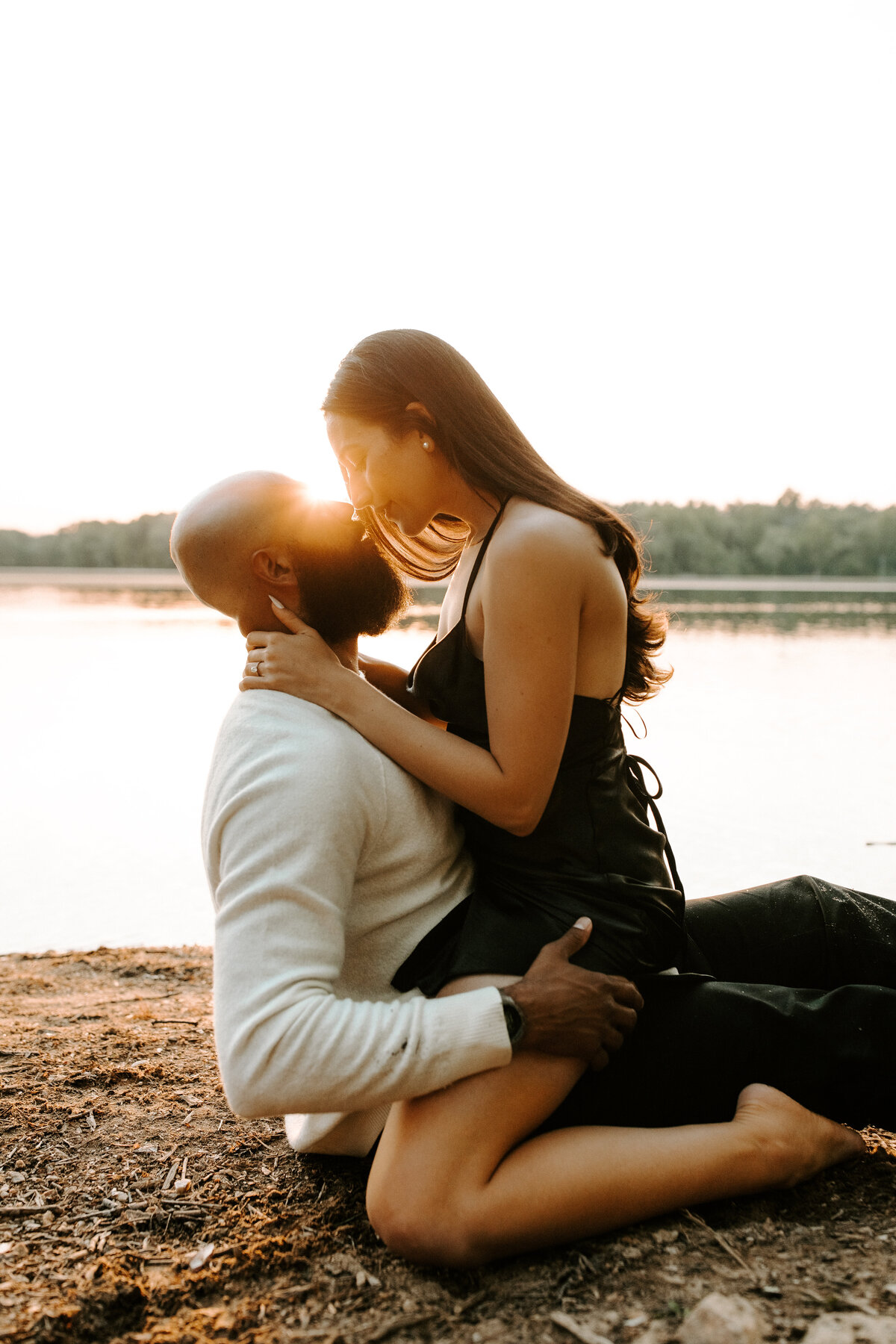 A male and female couple, the man is sitting on the ground while the woman straddles him. They are nose to nose as the sun sets right in between their faces.
