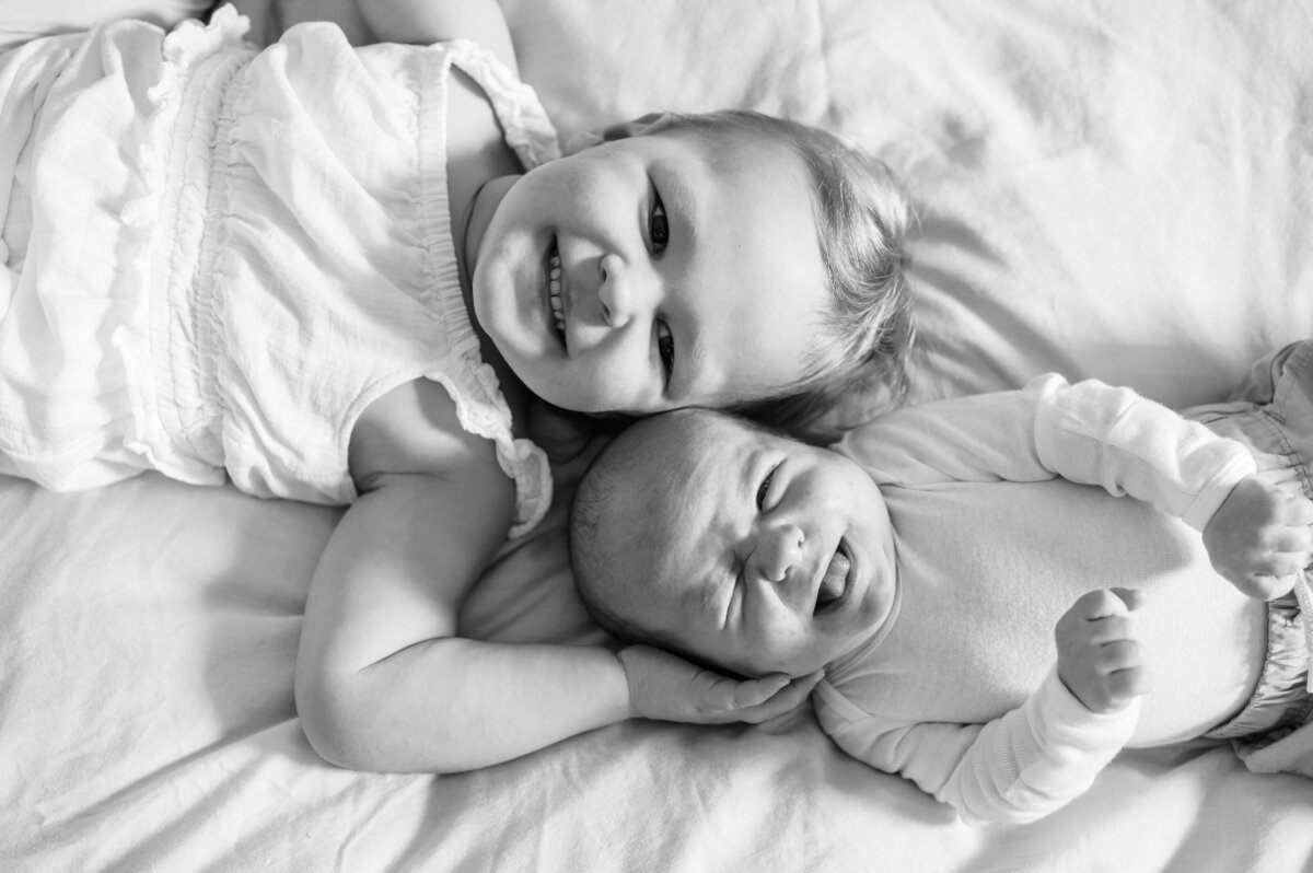 A black and white image shows a toddler daughter and her infant brother laying down on a bed with their faces together