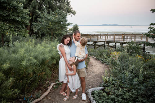 A Family of four look at eachother during their lifestyle family photos while standing on a trail at sunset after exploring around the Havre de Grace Promenade for a photoshoot.