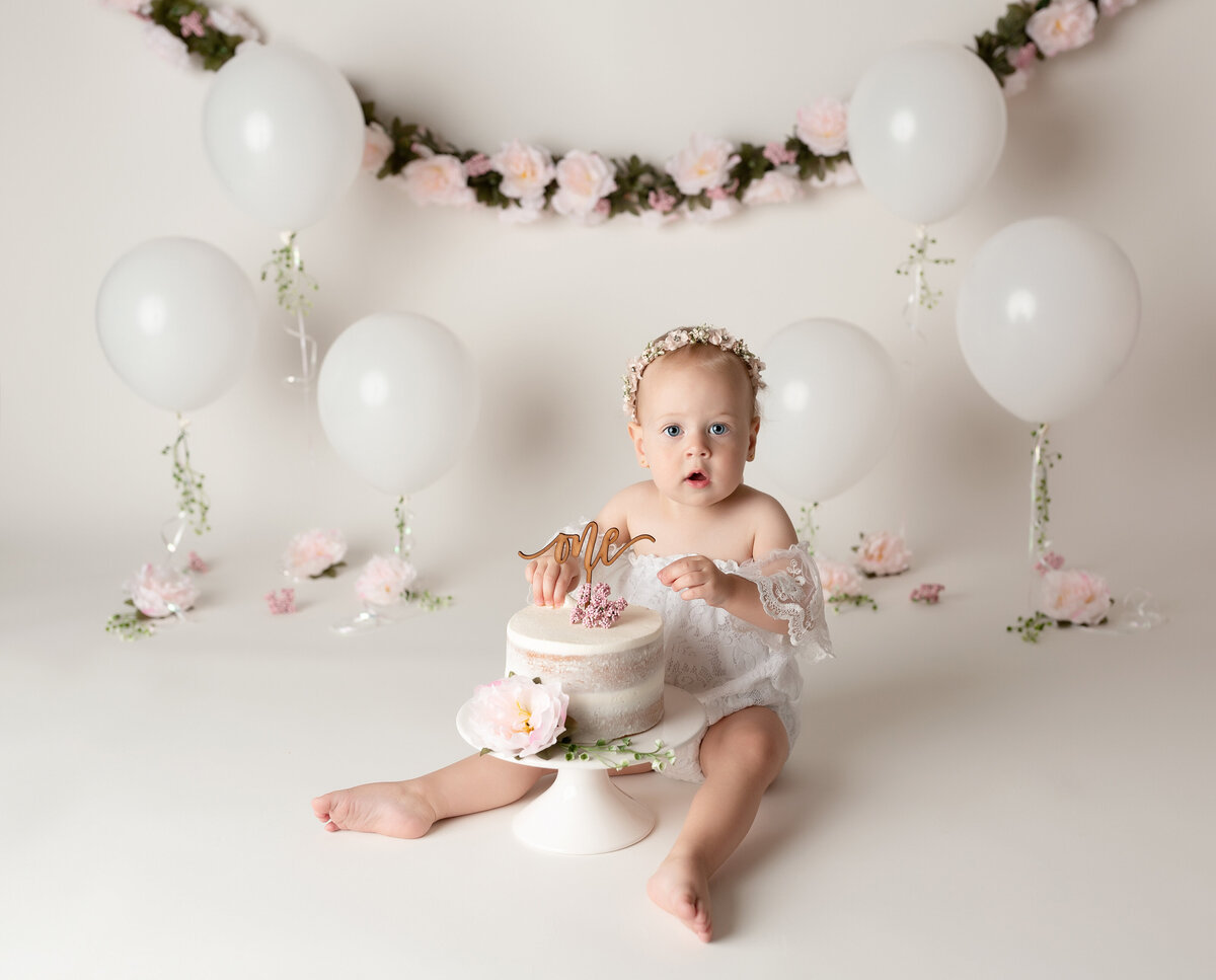 Elegant floral cake smash at West Palm Beach photography studio. Baby girl in a white lace romper is sitting with her cake between her legs looking at the camera and about to touch it for the first time. In the background, there are white balloons anchored to the ground with greenery and blooms.