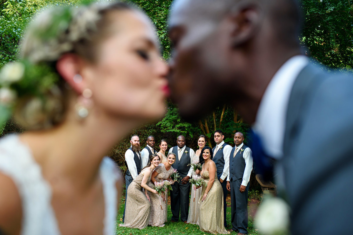 The wedding party in between the out of focus bride and groom kissing.