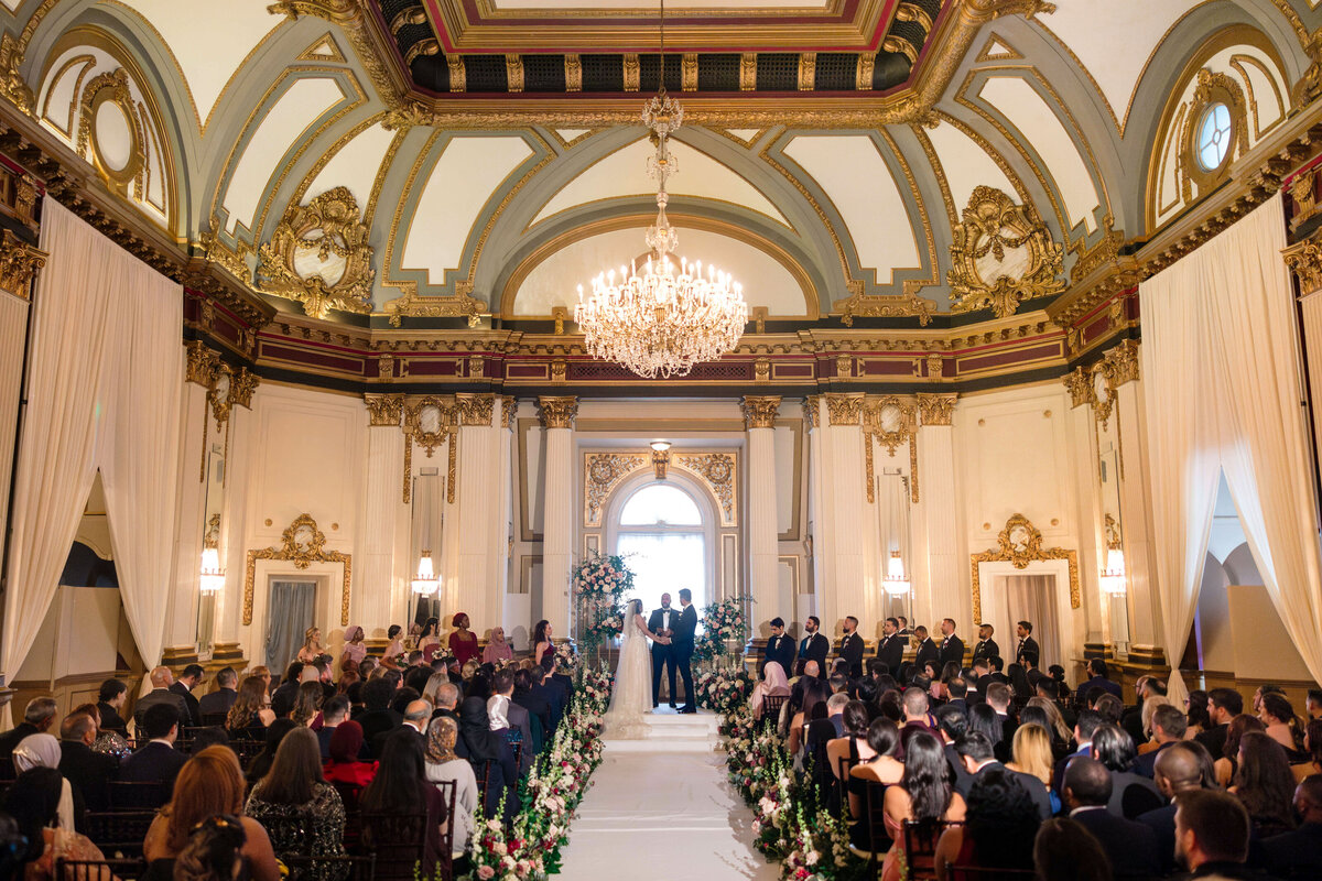 A grand indoor wedding ceremony in a lavish, ornate hall featuring high ceilings and a large chandelier. A couple stands at the altar with an arch of flowers, facing seated guests lining both aisles adorned with floral arrangements.