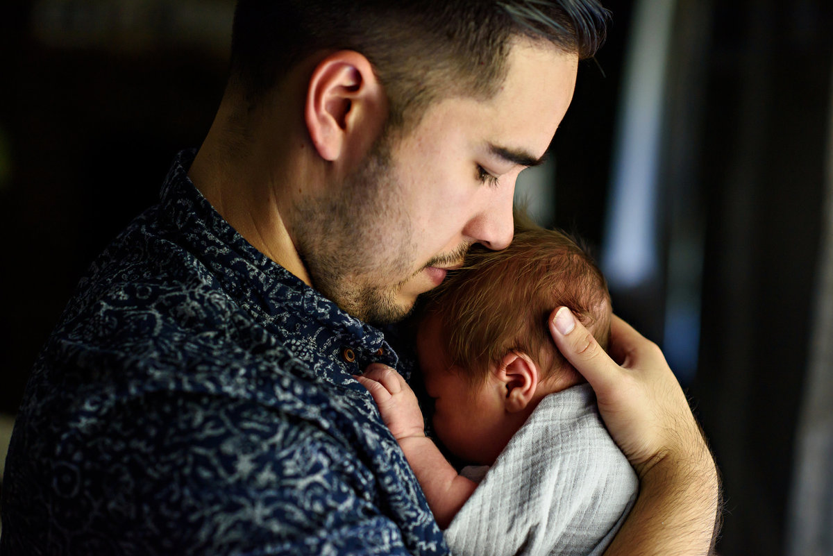A new father holds his week old baby to his chest and kisses his head.