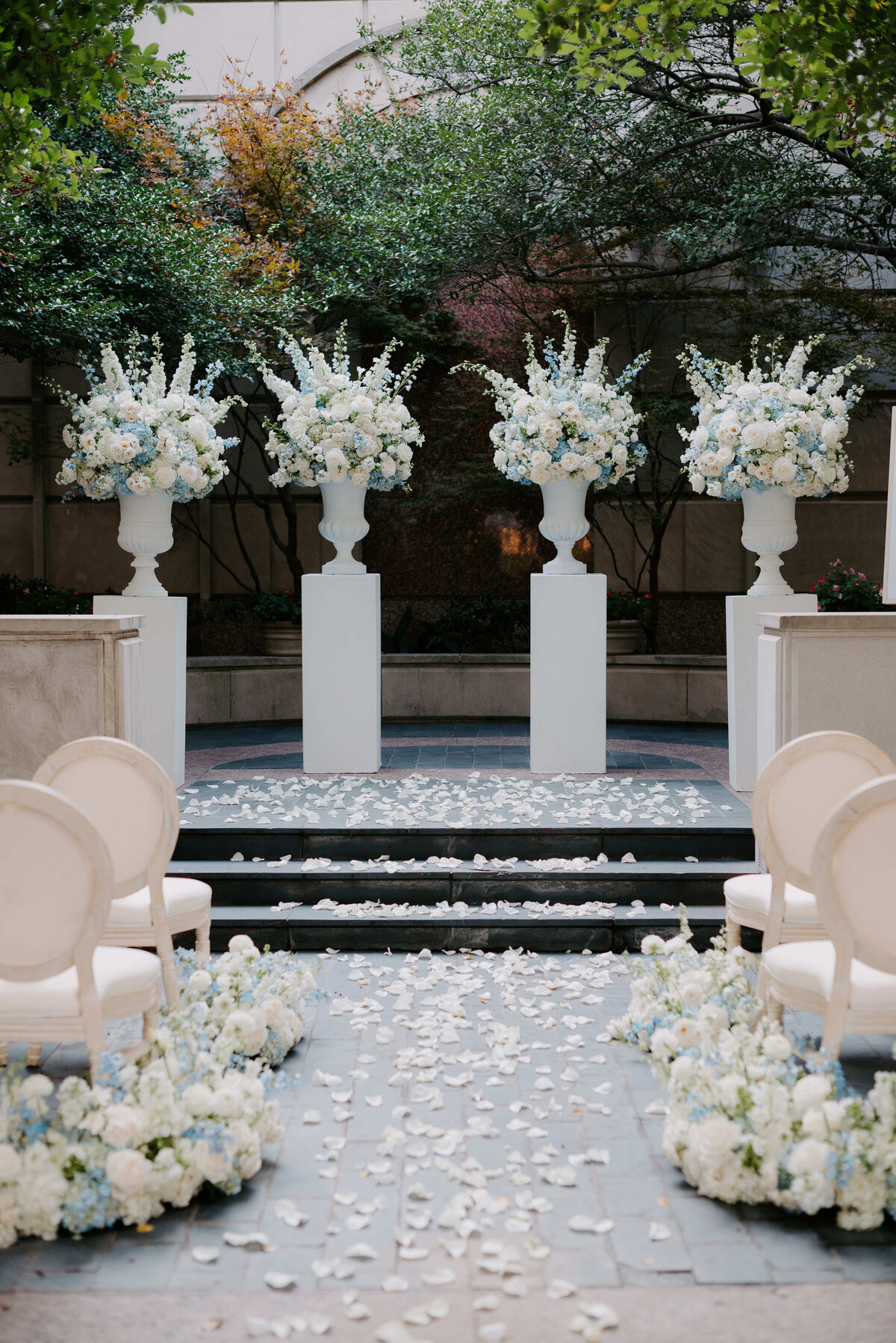 Elegant outdoor wedding ceremony setup with white chairs lining a flower-petal-strewn aisle. Tall vases of white and blue flowers on pedestals create a serene, romantic atmosphere under lush green trees.