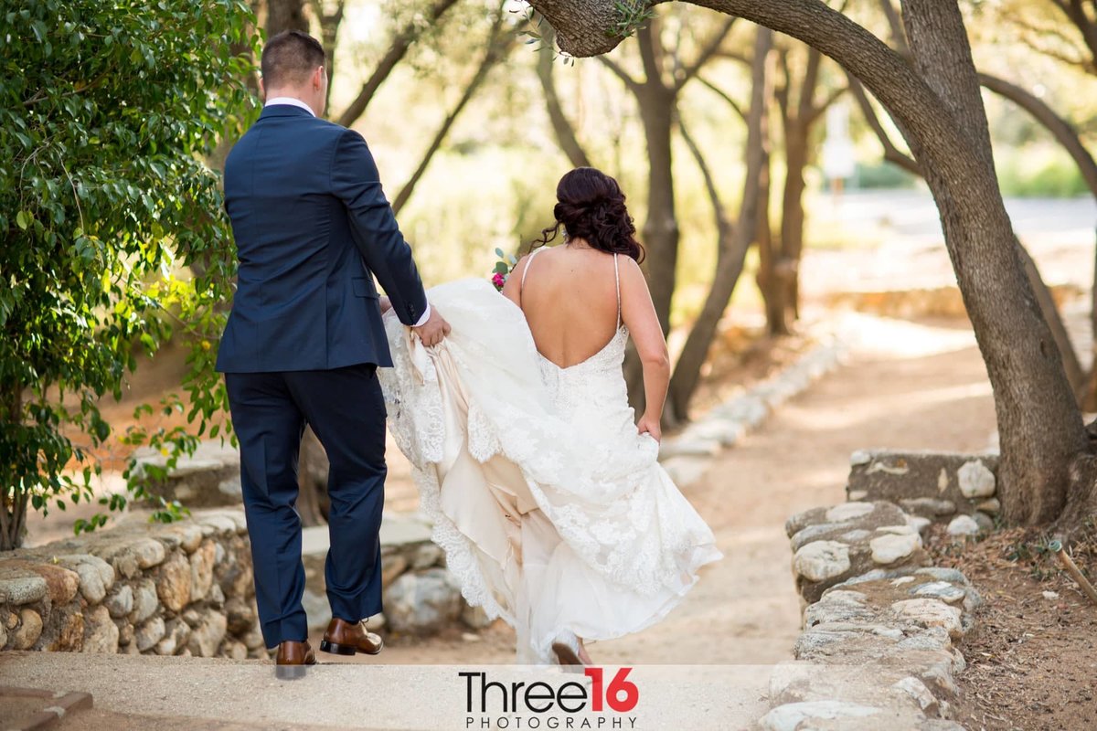 Bride and Groom walk down a dirt path while he holds her dress from touching the ground
