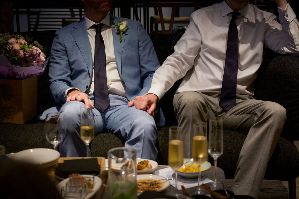 Grooms hold hands sat down.  Mercer wedding reception