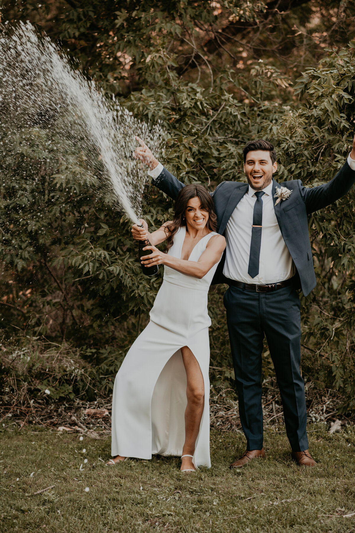 Bride and Groom pose for a fun and celebratory wedding photo after their backyard wedding ceremony in London, Ontario. Bride is spraying champagne in the air and the groom has his hands in the air, laughing. Captured by London, ON top wedding photographer Ashlee Ellison Photos and Films