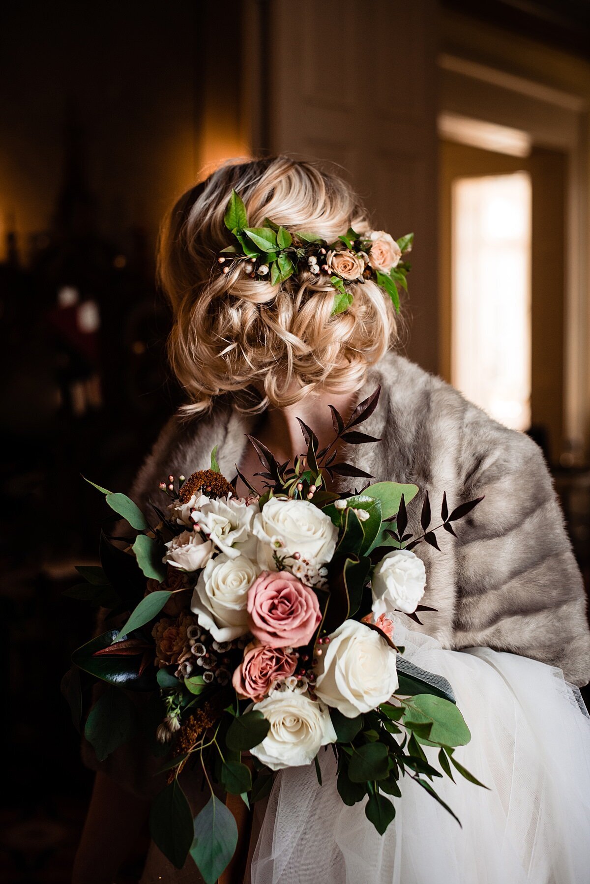 Detail of the bride's flowers. The bride has a floral hair comb in her blonde hair filled with greenery, blush flowers and purple berries.  The bride is wearing a fur wrap and is holding the tulle train of her skirt over her arm. In her hand is  a large bouquet of pink, blush and ivory roses with dark greenery accents.