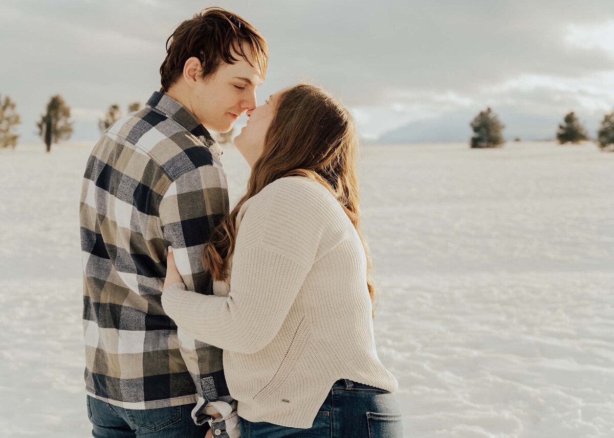 Maddie Rae Photography a couple standing close together giving eskimo kisses. he is facing away and she is facing the camera. it's snowy and there are mountains in the background