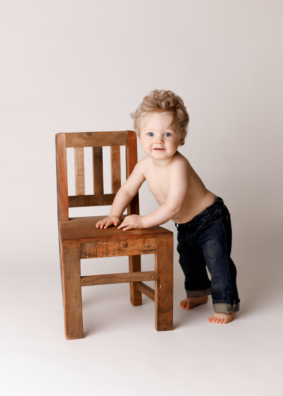 One-year milestone photoshoot in West Palm Beach, FL photo studio. Baby boy with curly blonde hair wearing jeans is standing up against a miniature chair.
