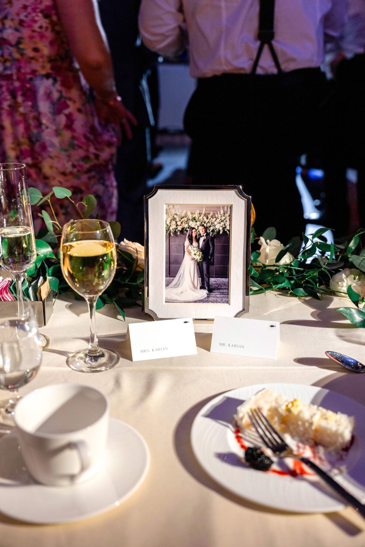 A wedding reception table with a framed photo of a couple, labeled seats for Mr. and Mrs. Lanza, a slice of cake with berries on the plate, a cup, and glasses of wine and champagne. Floral decor surrounds the setting.